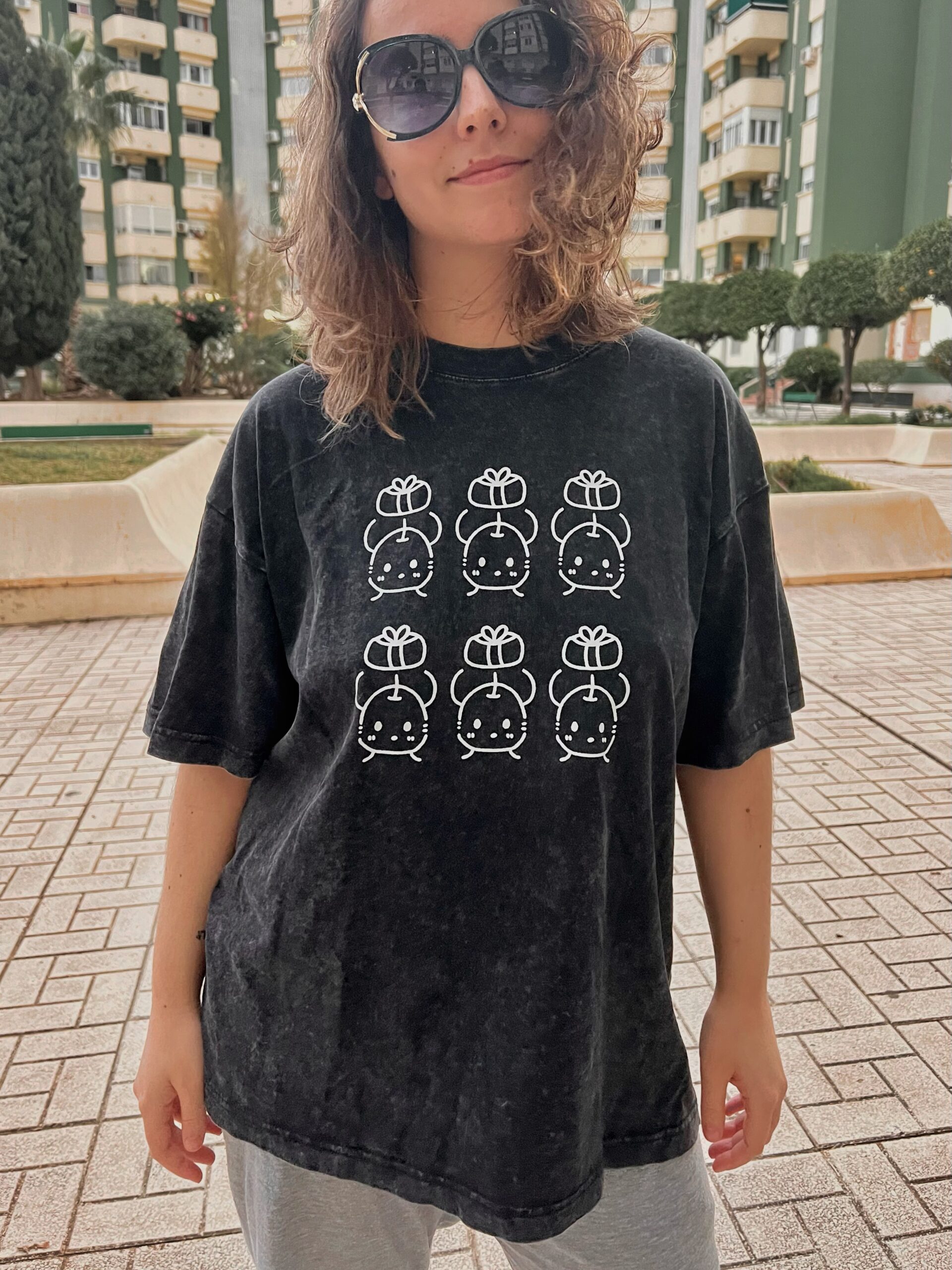 Camisetas de junimos sosteniendo regalos. Diseño en blanco. Los colores de la camiseta pueden ser en negro o en morado. Los modelos son una mujer con la camiseta negra y un hombre con la camiseta morada