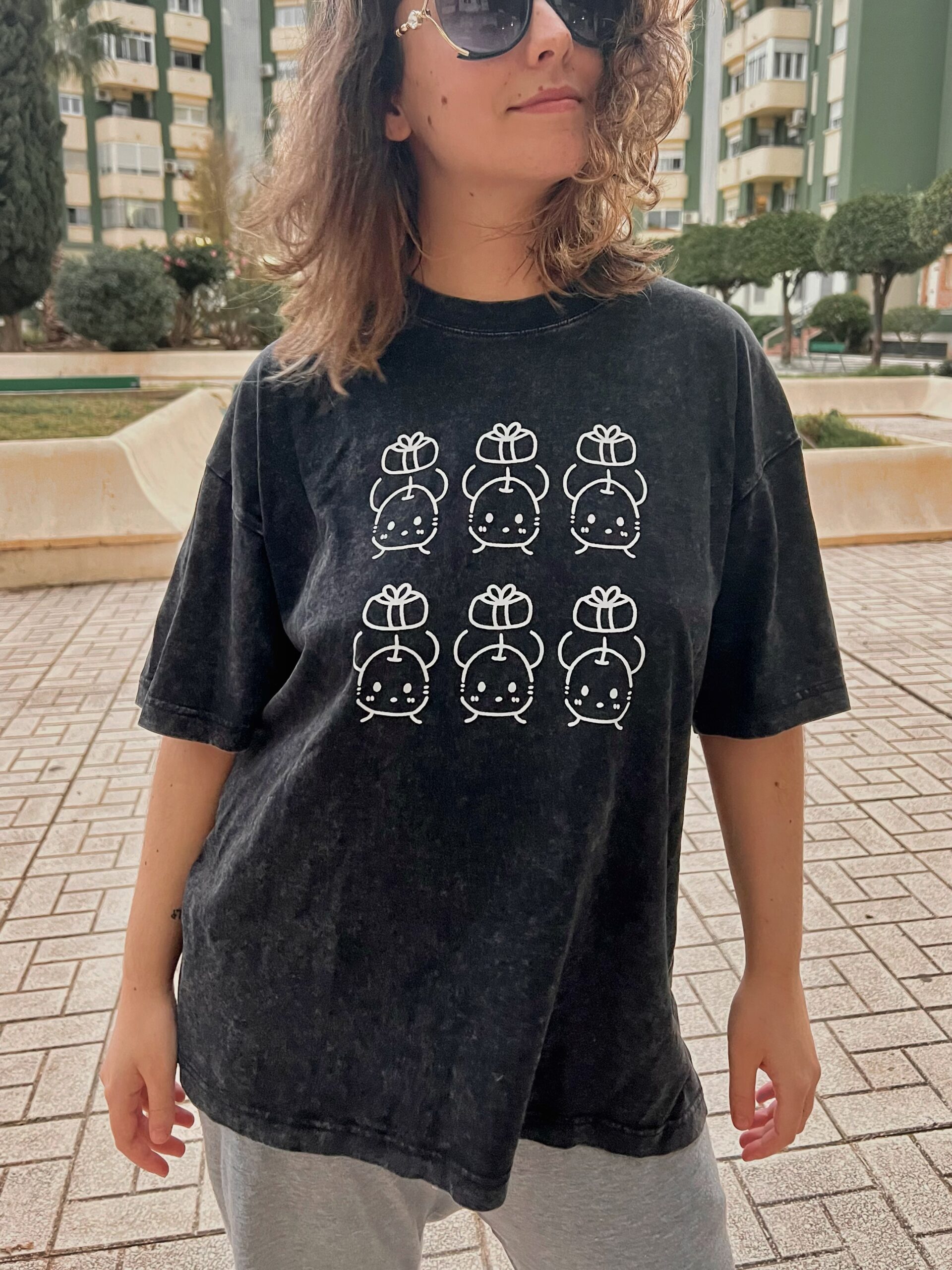 Camisetas de junimos sosteniendo regalos. Diseño en blanco. Los colores de la camiseta pueden ser en negro o en morado. Los modelos son una mujer con la camiseta negra y un hombre con la camiseta morada