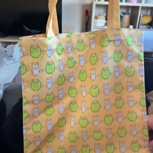 En esta tote naranja tirando a amarillo pastel se ve a Benjamín, el gato aceituno, con y sin su aceituna en un diseño estilo mosaico. Benjamín es uno de mis diseños propios y el icono de Mochopo. Es un gatito al que le gusta dormir dentro de una aceituna.