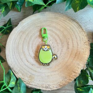 Llavero del gato aceituno o Benjamín, icono de Mochopo. Acabado de resina epoxi. Verde con mosquetón verde también. Fondo de madera y hojas verdes.