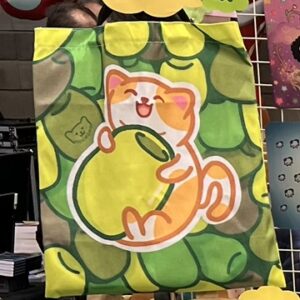 Una tote bag de Benjamín, el gato aceituno, abrazado a su aceituna con un fondo de diferentes tipos de aceitunas