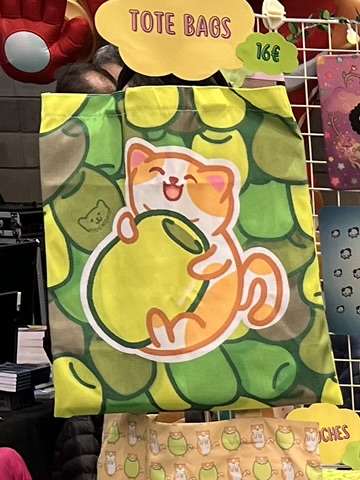 Una tote bag de Benjamín, el gato aceituno, abrazado a su aceituna con un fondo de diferentes tipos de aceitunas