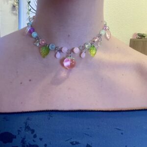 Collar artesanal con un melocotón de vidrio en el centro. Tiene cuentas de vidrio rosa y verdes así como gotas rosas y hojas verdes que cuelgan