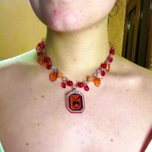 Collar stardew valley con cuentas de vidrio rojo y gotas y hojas rojas que cuelgan. En el centro tiene un charm acrílico de semilla de fresa del videojuego stardew valley.