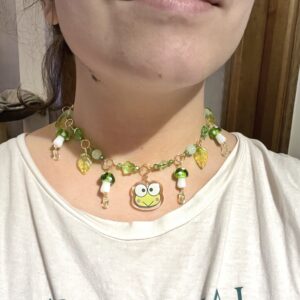 Collar Sanrio con un Keroppi (rana verde) en el centro. Metal de acero inoxidable con baño de oro del que cuelgan abalorios y setas verdes