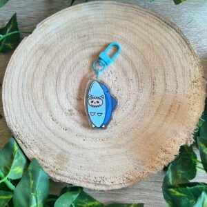 Llavero del gato tiburón, un gato o gata naranja crema con un traje de tiburón. Acabado de resina epoxi. Llavero de mosquetón azul. Fondo de madera y hojas verdes.