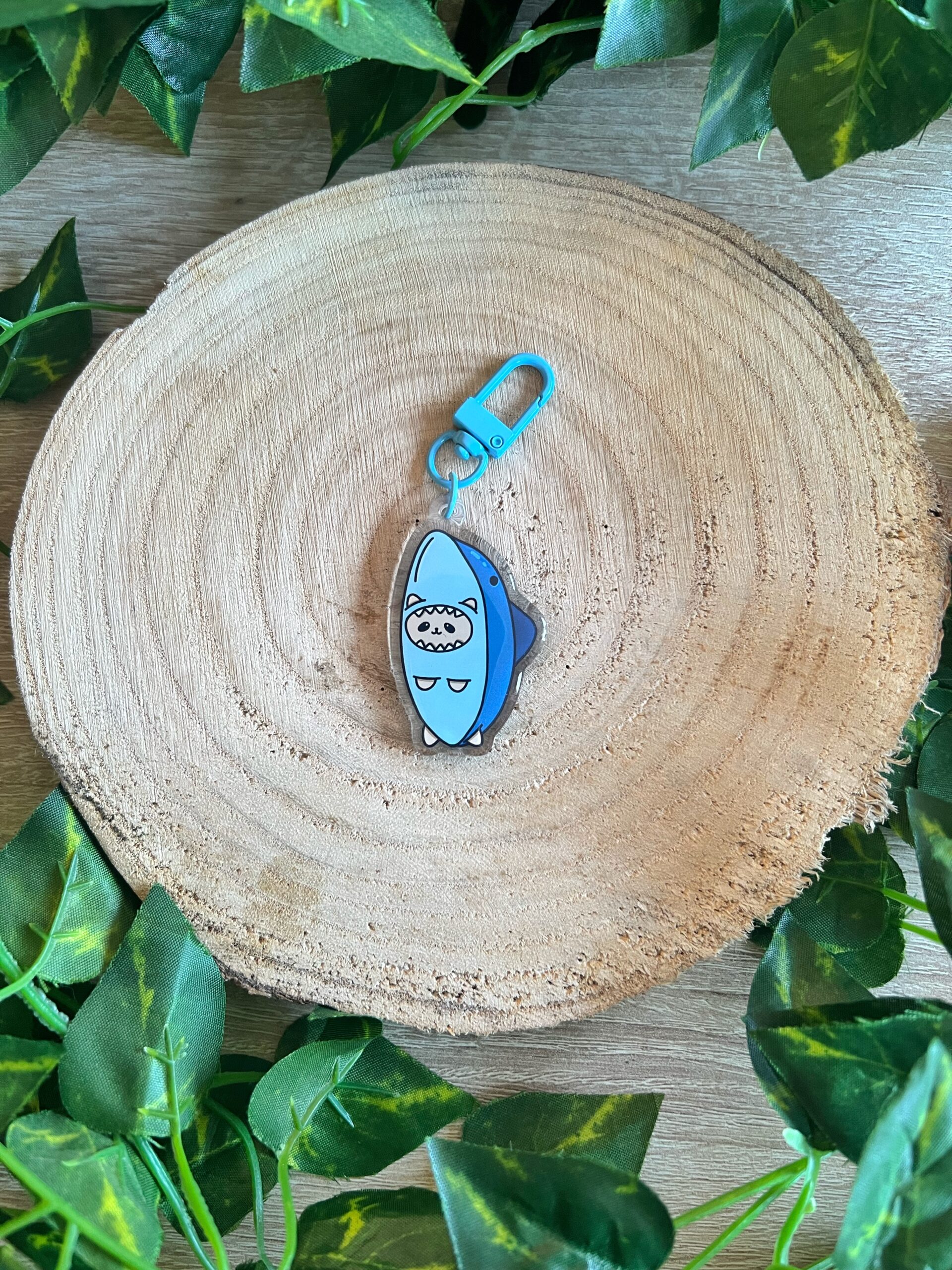 Llavero del gato tiburón, un gato o gata naranja crema con un traje de tiburón. Acabado de resina epoxi. Llavero de mosquetón azul. Fondo de madera y hojas verdes.