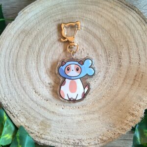 Llavero del gato pez, un gato o gata tricolor con un gorro de pez. Acabado de resina epoxi. Llavero de mosquetón en forma de gato. Fondo de madera y hojas verdes.