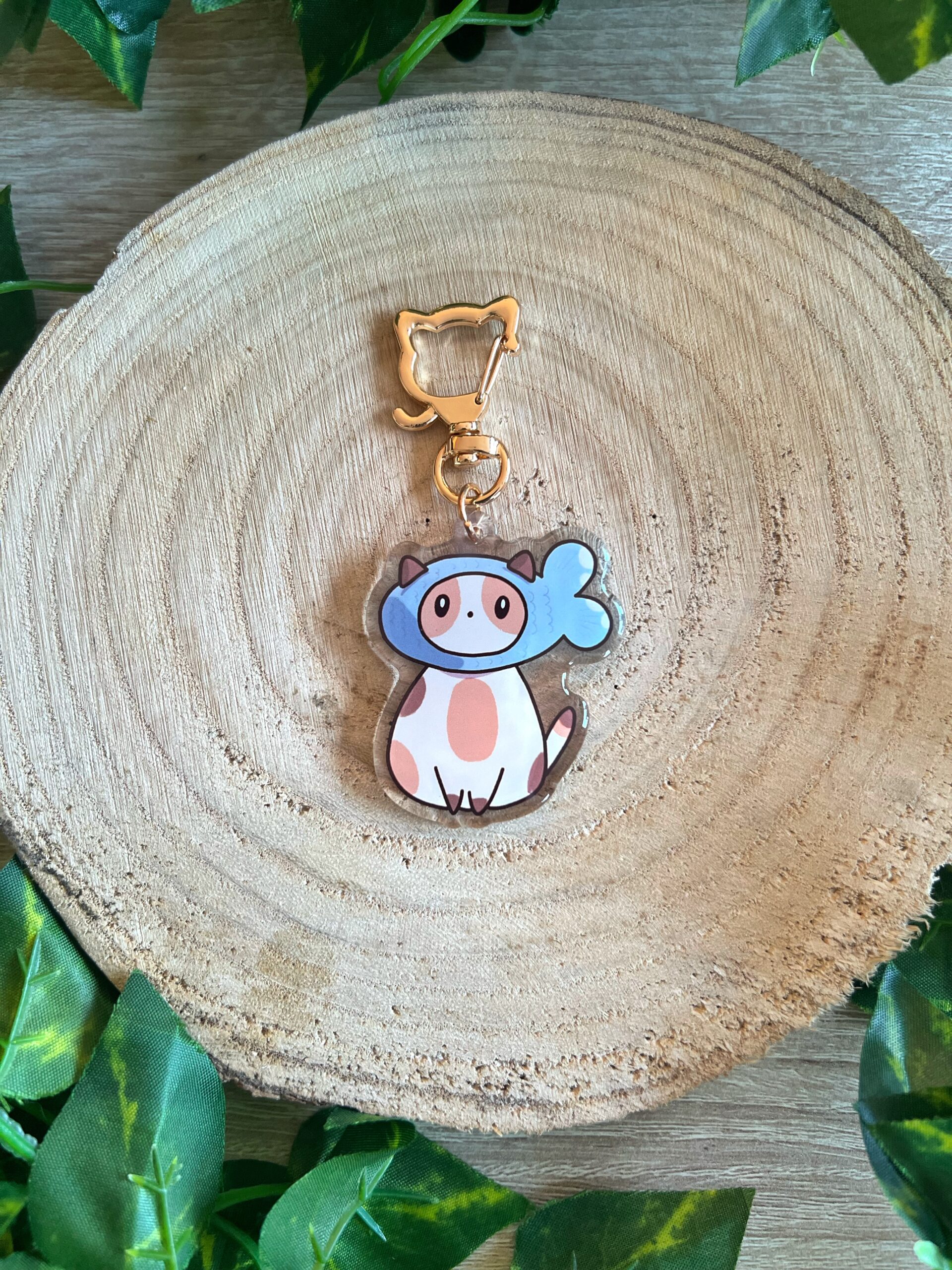 Llavero del gato pez, un gato o gata tricolor con un gorro de pez. Acabado de resina epoxi. Llavero de mosquetón en forma de gato. Fondo de madera y hojas verdes.