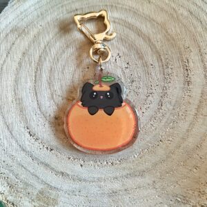 Llavero de Peluso el gato mandarino. Un gato negro de pelo largo metido en una mandarina naranja con una hojita en su cabeza. Llavero de mosquetón con forma de gato. Fondo de la imagen con hojas verdes y un trozo de madera